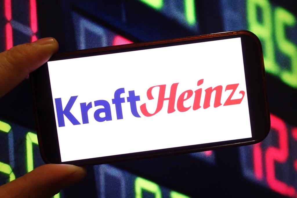 A hand holding a smartphone featuring the KRAFT Heinz company logo, symbolizing their predicted release of additional refreshing aromas of crystal light vodka in 2026.