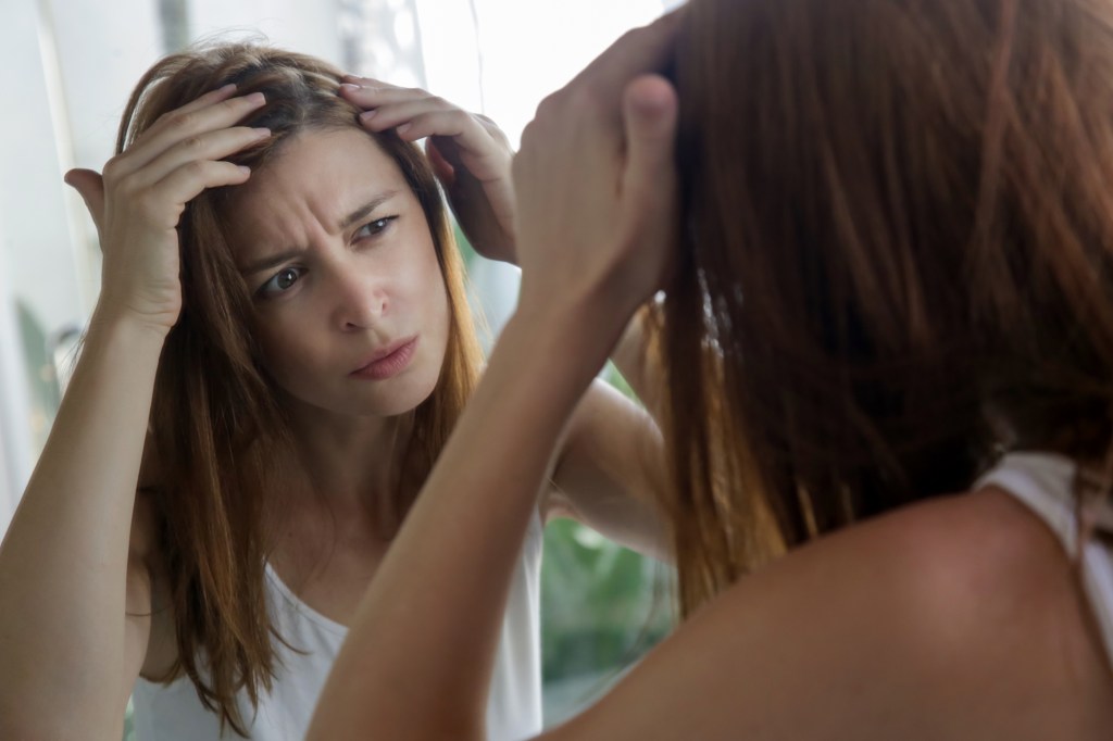 Young woman who examines her head and hair for gray strands and possible hair loss in front of a mirror