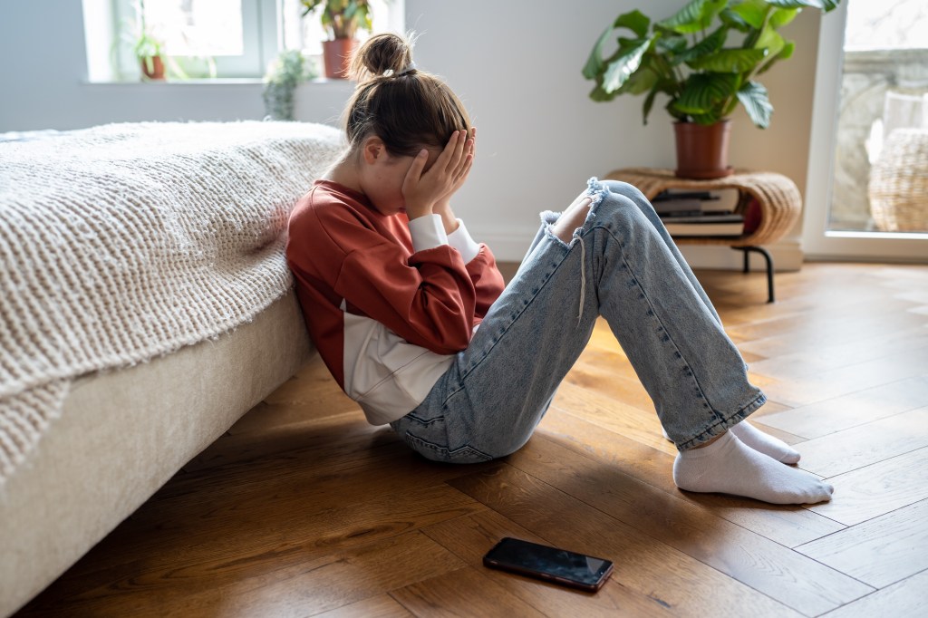 Dissatisfied teenage girl sitting on the floor, covering her face with her hands and crying with a nearby cell phone, representing the internet impact on the mental health of General Z