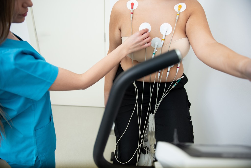 Thick woman undergoing cardiovascular fitness evaluation in a routine, monitored by a health care professional with electrodes attached to her chest